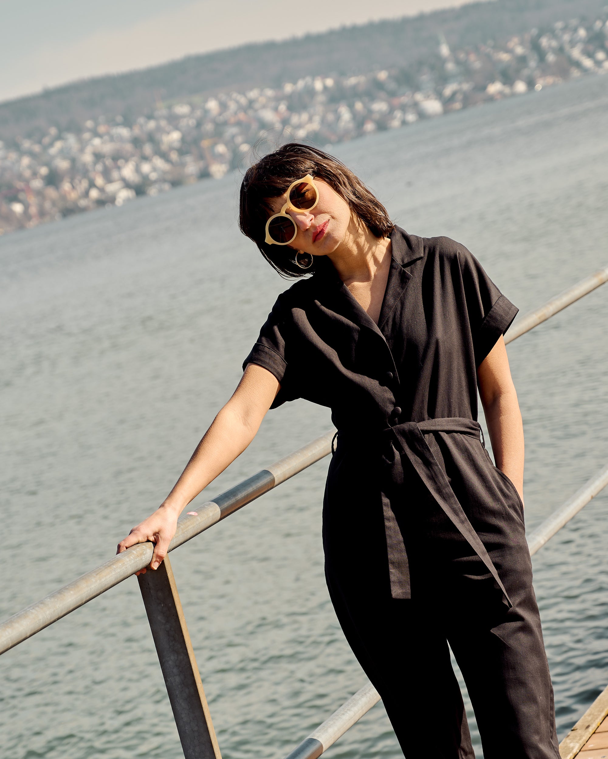 Petite model wearing the Black Jumpsuit while leaning on a railing by the lakeside.