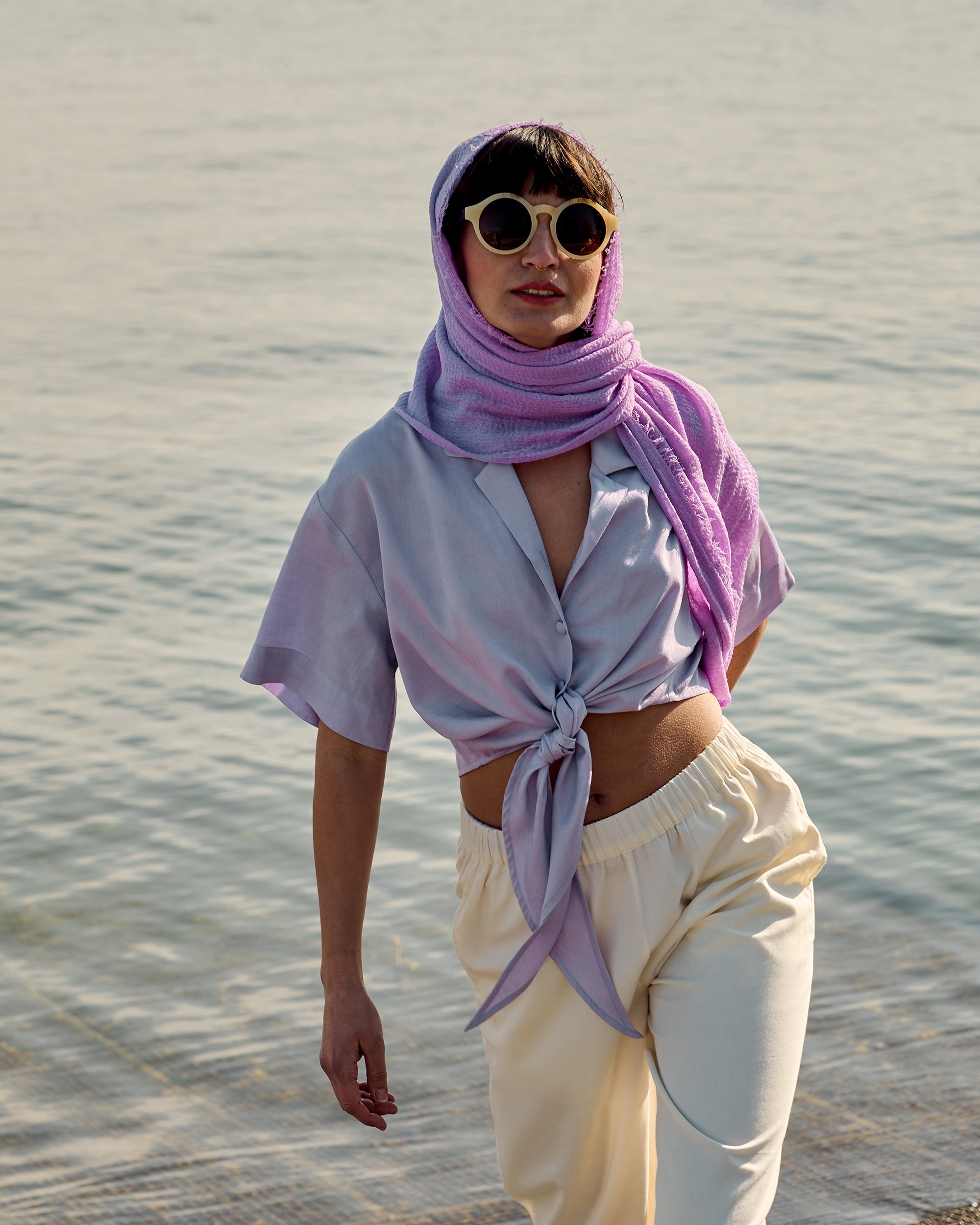 Petite model wearing the Wrap Blouse Lilac with cream wide-leg trousers by the lakeside.