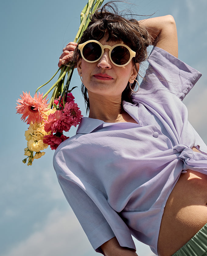 Petite model wearing the Wrap Blouse Lilac with sunglasses, holding flowers against a blue summer sky.