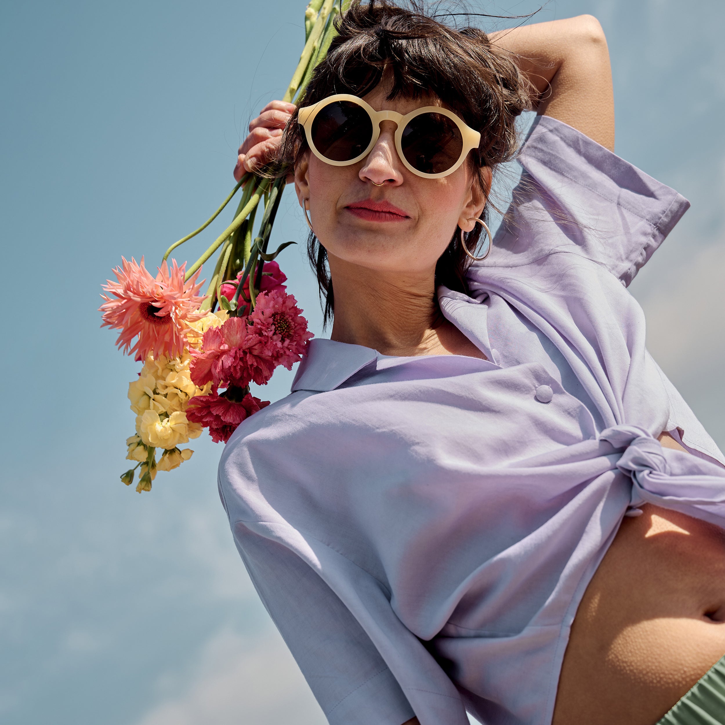 Petite model wearing the Wrap Blouse Lilac with sunglasses, holding flowers against a blue summer sky.