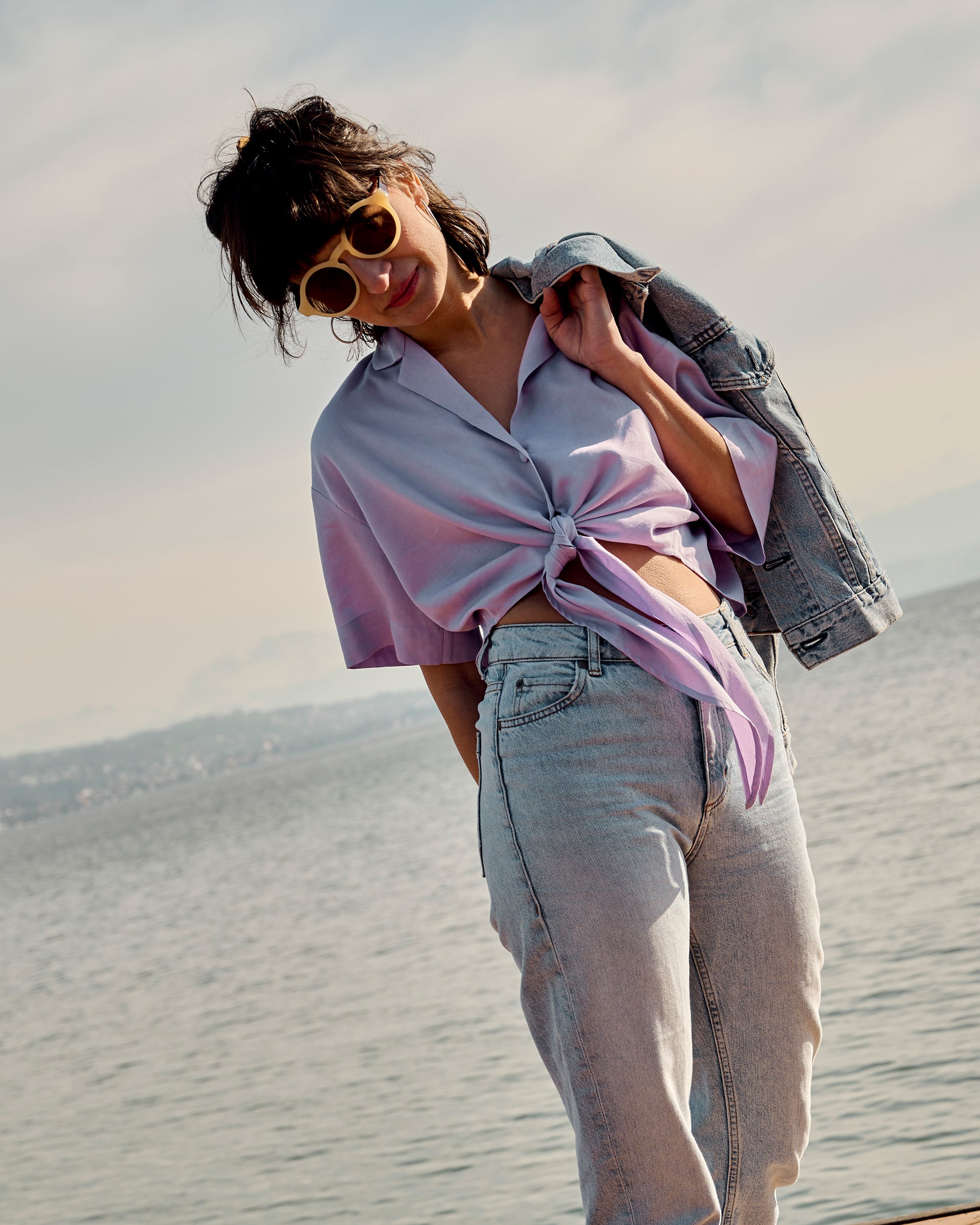 Petite model wearing the Wrap Blouse Lilac with a denim jacket over the shoulder by the lakeside.
