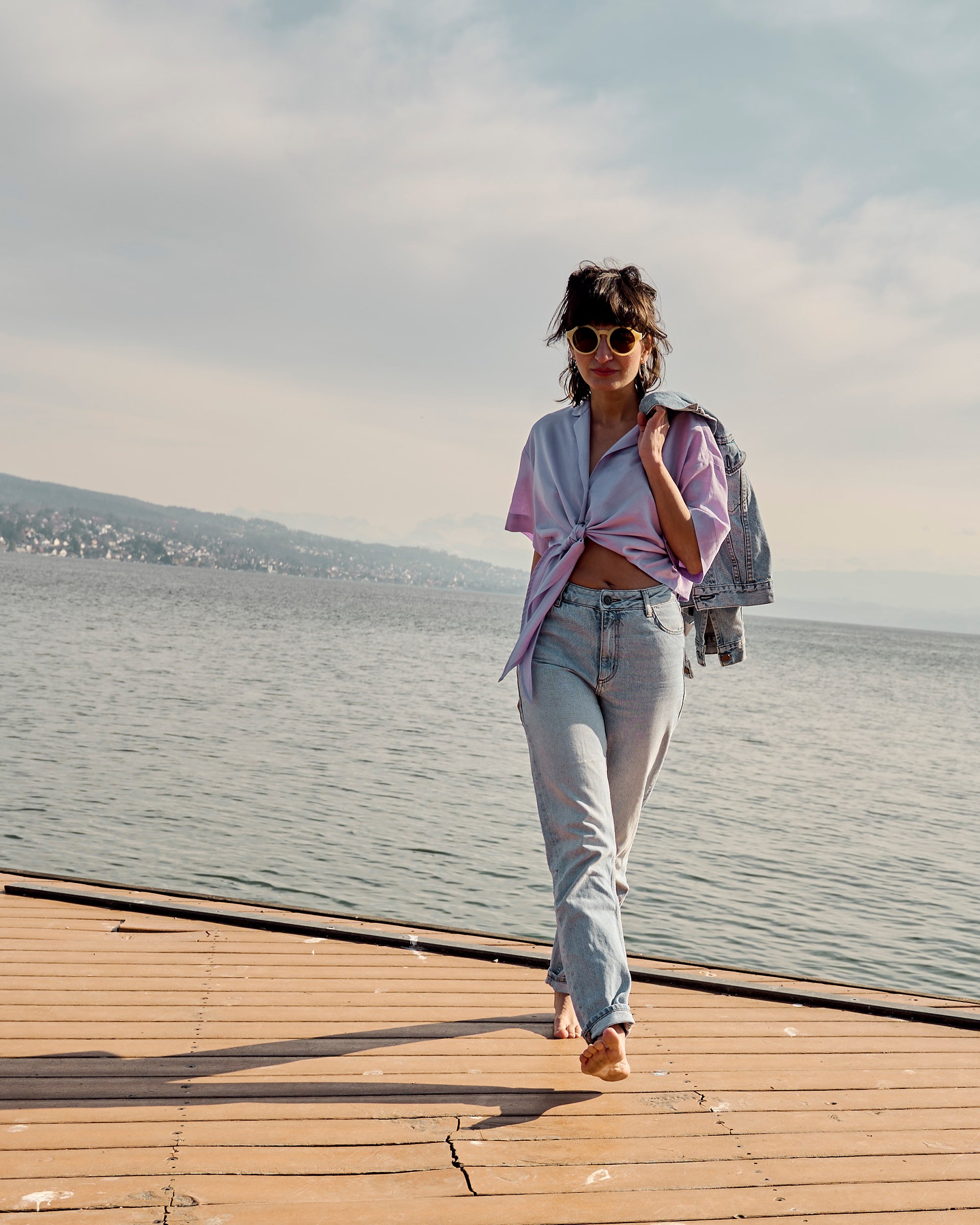 Petite model wearing the Wrap Blouse Lilac and light denim jeans with a denim jacket over the shoulder by the lakeside.